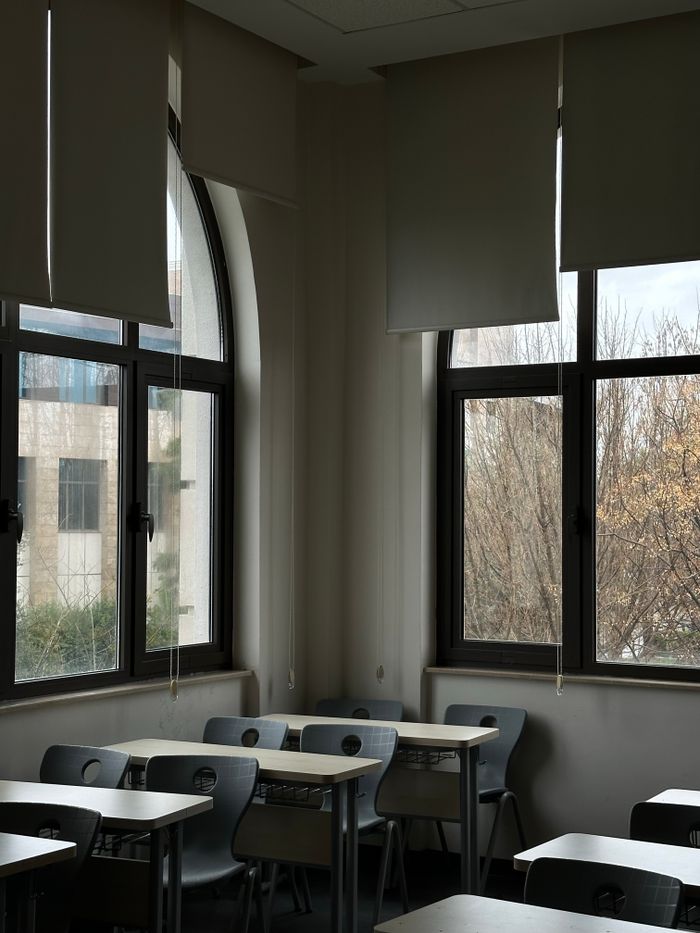 Sunlit empty classroom with desks and chairs, featuring large windows with blinds.